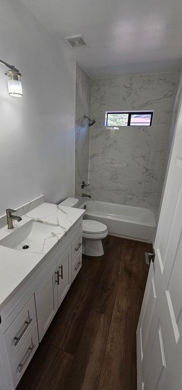 Narrow white bathroom with vanity, toilet, tub, dark wood floor, and small window above the shower