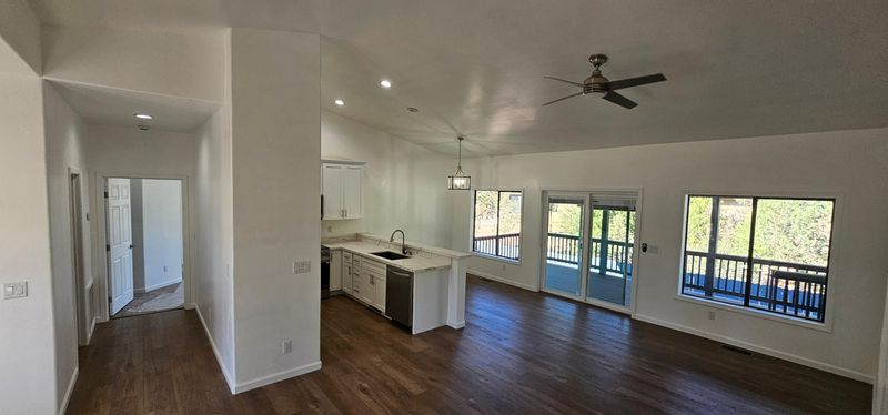 Bright empty living room with kitchen, dark wood floors, ceiling fan, and large windows opening to a balcony