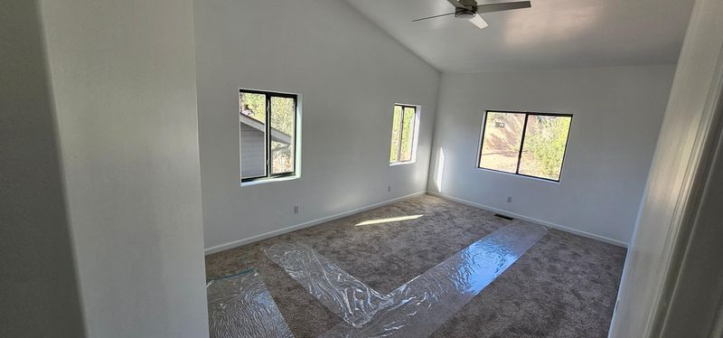 Empty white room with carpet, three windows, and a ceiling fan, lit by sunlight