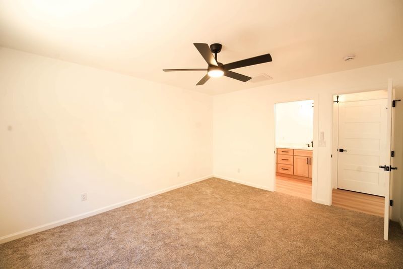 Empty carpeted bedroom with beige walls, ceiling fan, and an open doorway to a bathroom vanity