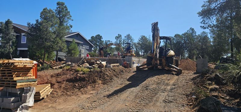 Construction site with excavator grading a dirt lot beside houses under a clear blue sky