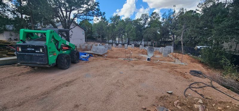 Green construction loader on a dirt lot with materials and trees under a cloudy sky