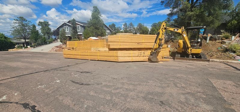 Yellow lumber stacked beside a yellow excavator on a paved lot under a blue sky