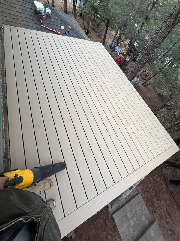 Worker installing beige roof panels on a sloped roof, with trees and a brick wall nearby