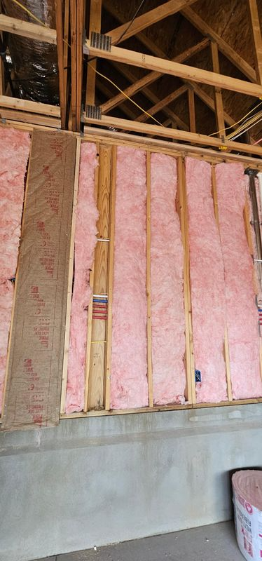 Unfinished wall framing with pink insulation batts and exposed wooden roof trusses above a concrete foundation.