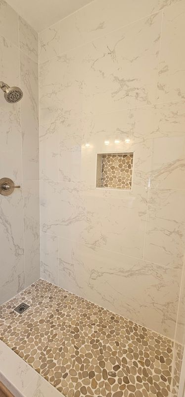 Bathroom shower with white marble-look tile and pebble floor, plus built-in niche shelf.