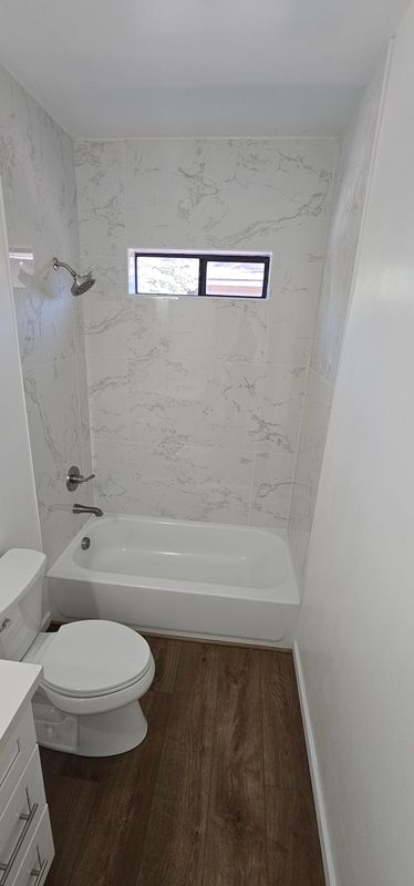 Small white bathroom with a tub-shower, toilet, wood-look floor, and a narrow window.