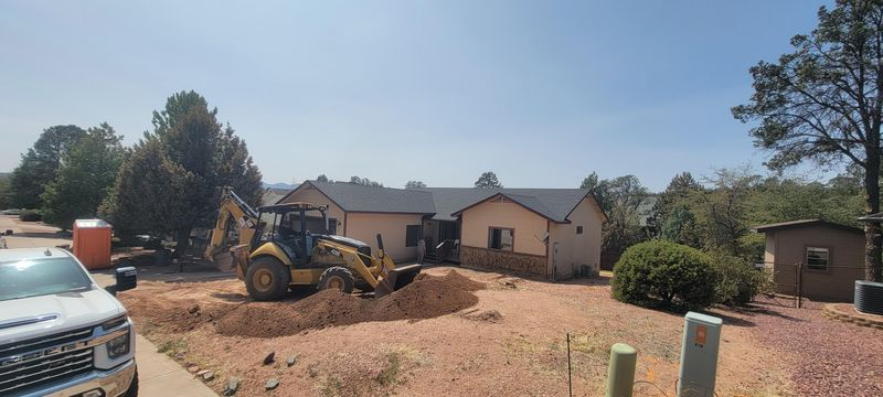 Construction equipment leveling a dirt yard in front of a beige house under a clear blue sky