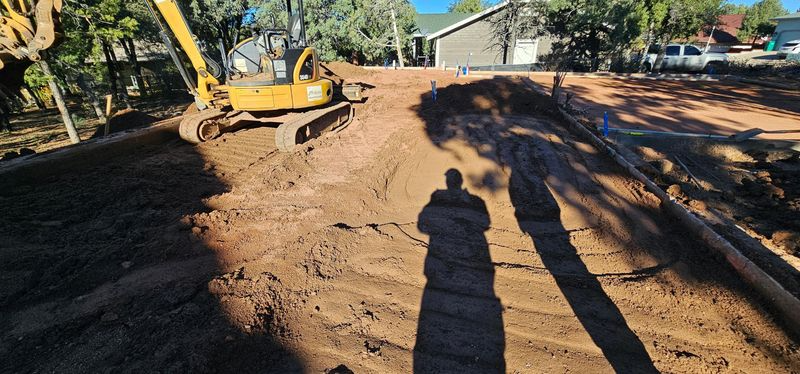 Construction site with excavator leveling a dirt lot beside trees and houses in bright sunlight
