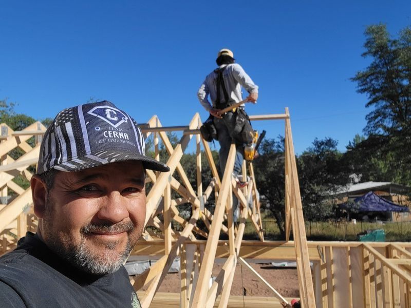 Man in cap taking selfie at a wooden house frame, with a worker on scaffolding behind him under blue sky