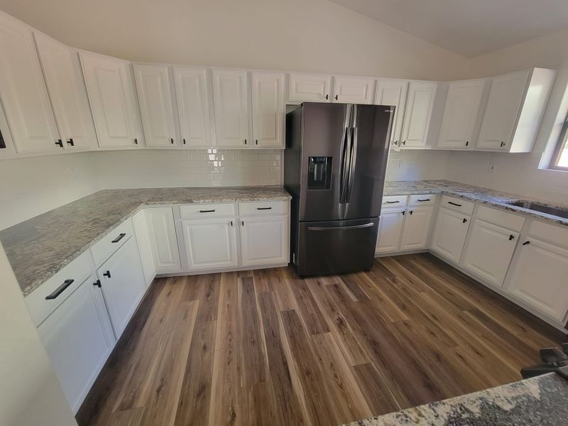Bright kitchen with white cabinets, gray countertops, stainless fridge, and wood floors.