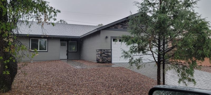 Gray house with a white garage door, gravel driveway, and trees under overcast skies.