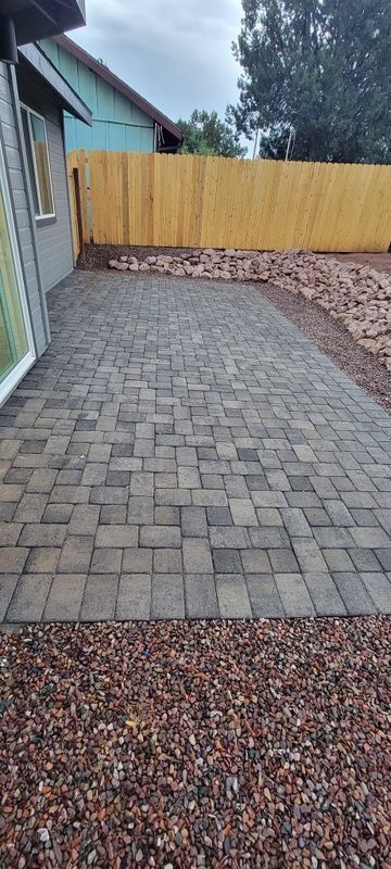 Backyard patio of gray pavers bordered by red gravel, with a wooden fence and house wall.