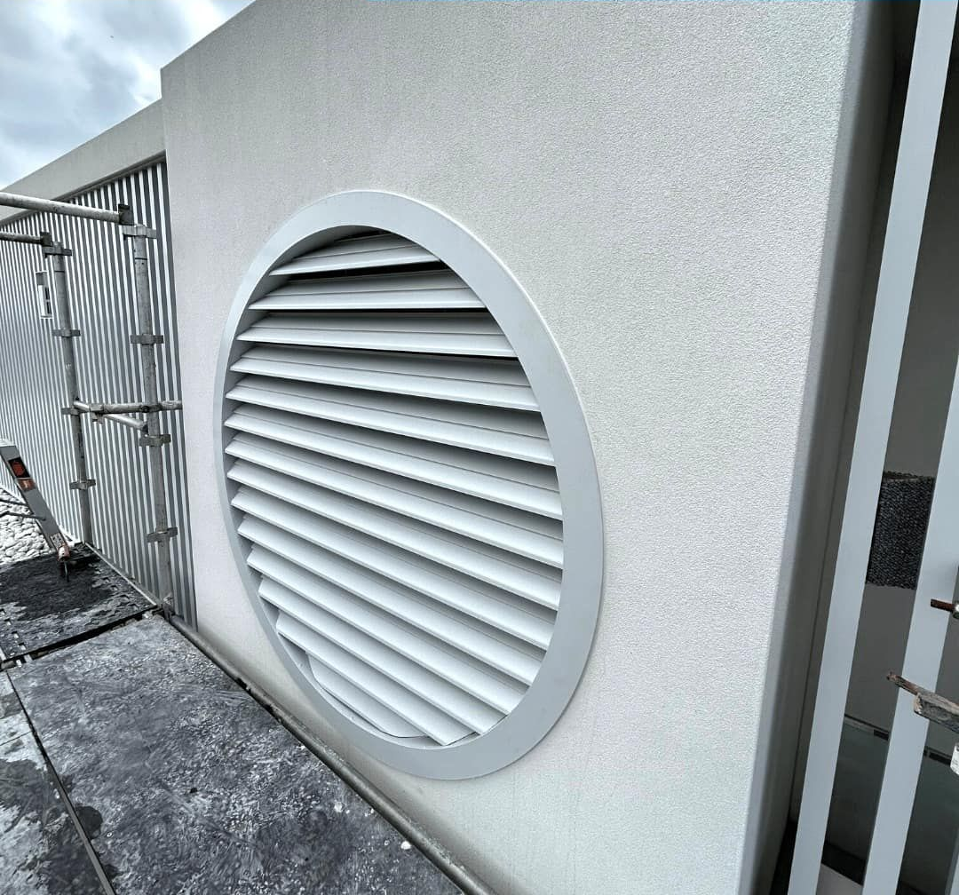 A round, silver aluminium louvre vent installed on a textured, light-coloured concrete exterior wall — Highlander Metals in Kirrawee, NSW