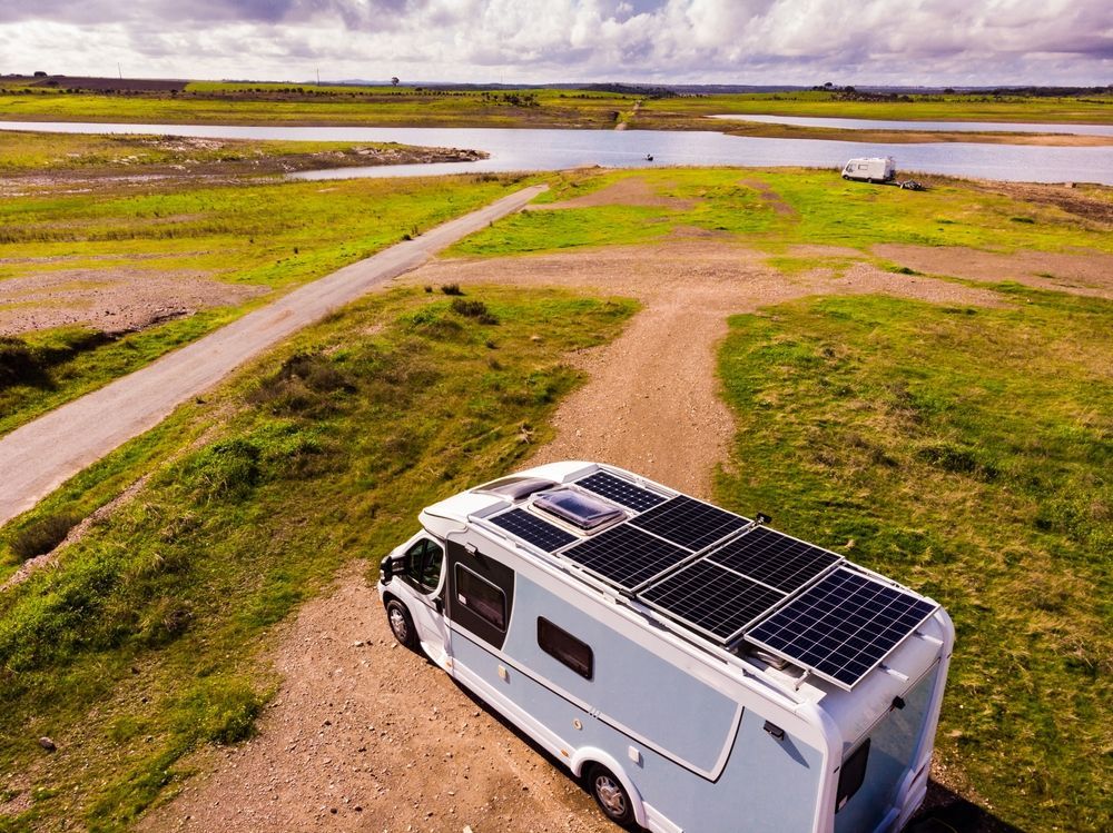 RV parked in field with solar panels on roof, overlooking water under cloudy sky.