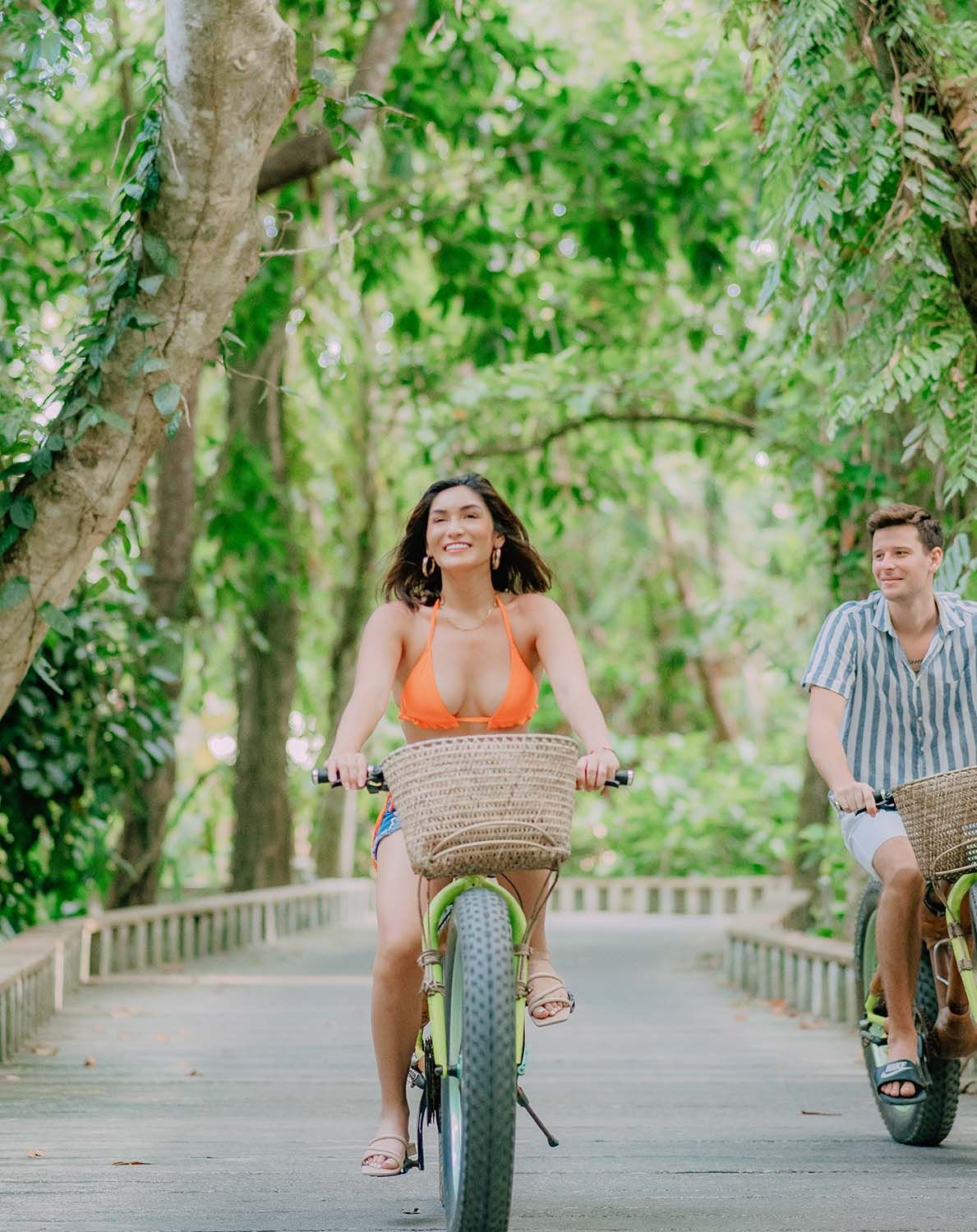 Two people riding bicycles on a tree-lined path, one in front with a basket on the bike.