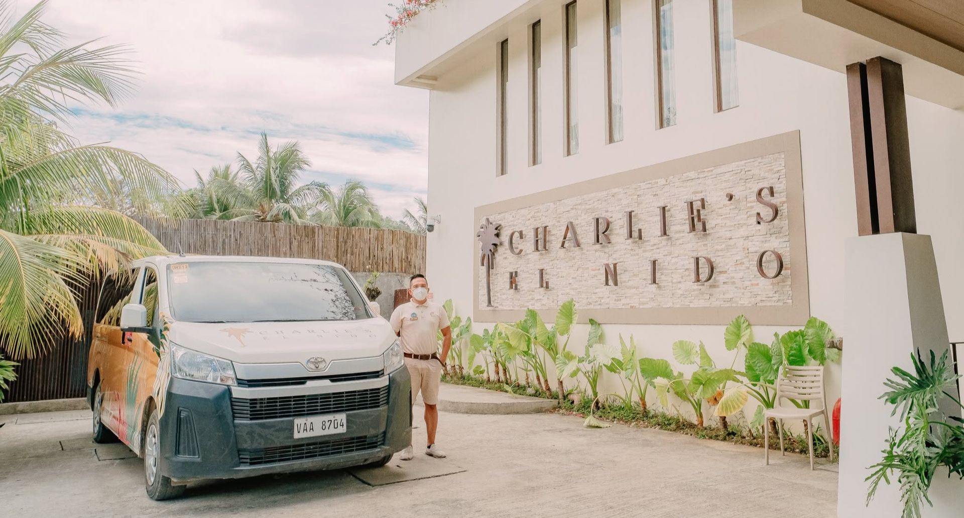 Van parked outside a modern building with a large sign and tropical plants along the entrance.