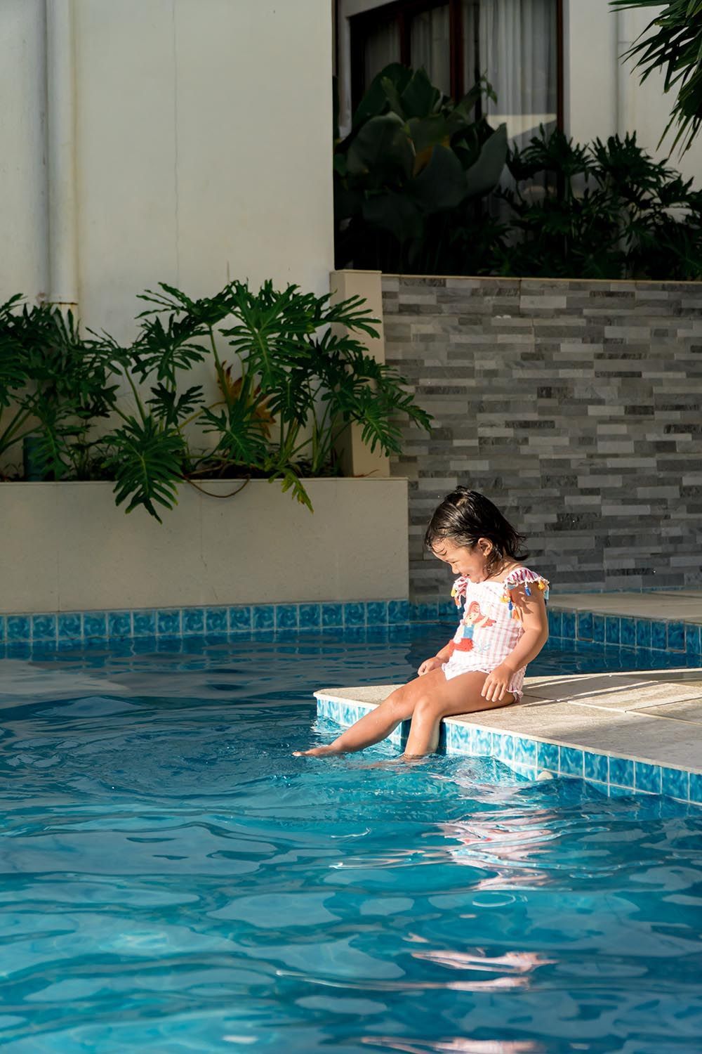 Child sitting on pool edge with feet in water, beside a blue swimming pool and green plants.