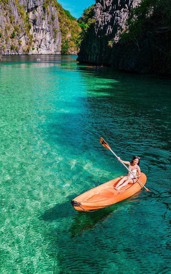 Person kayaking on turquoise water beside steep rocky cliffs and a lush island in El Nido