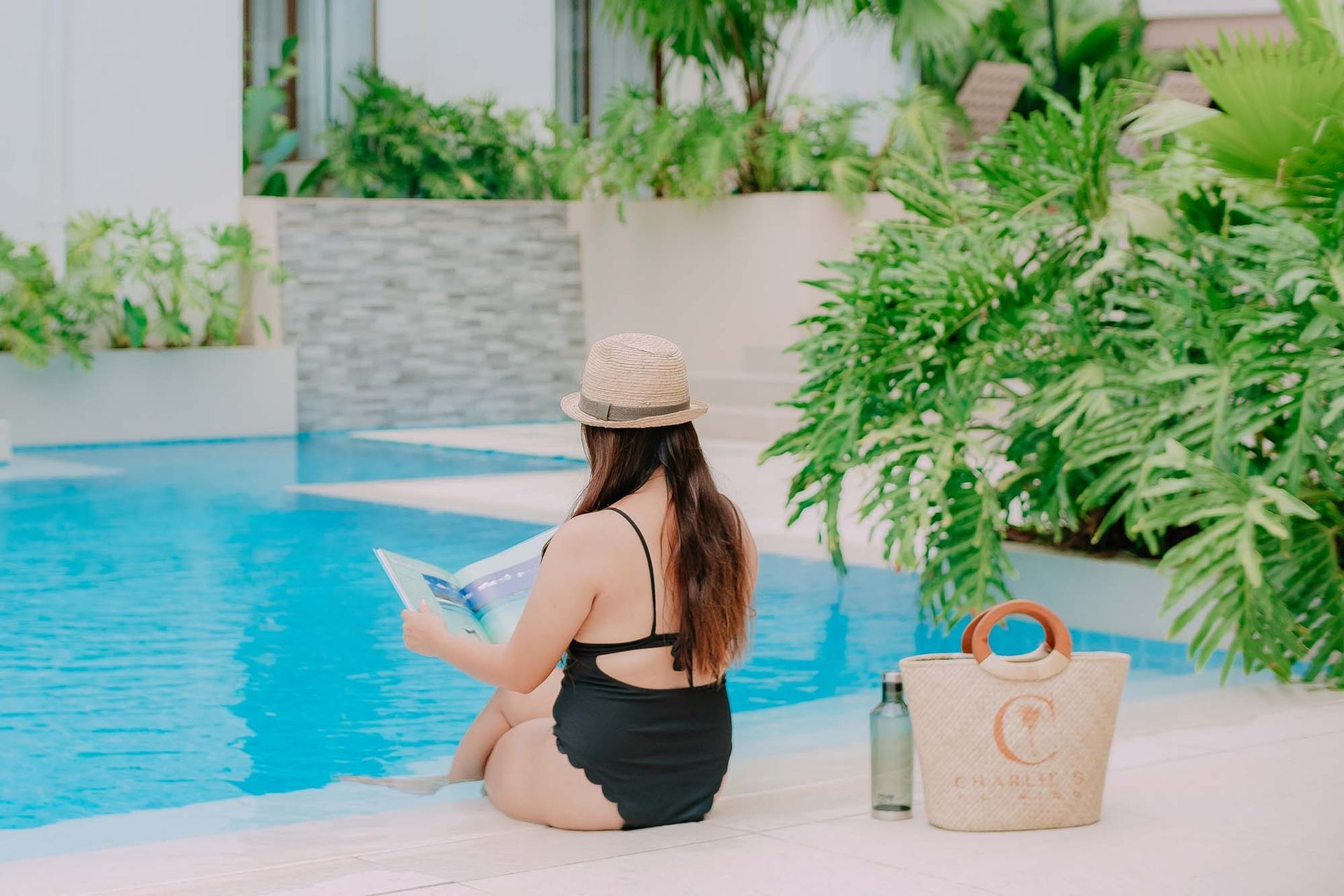Woman in black swimsuit and straw hat sits by a pool, reading a book. Bag and bottle beside her.