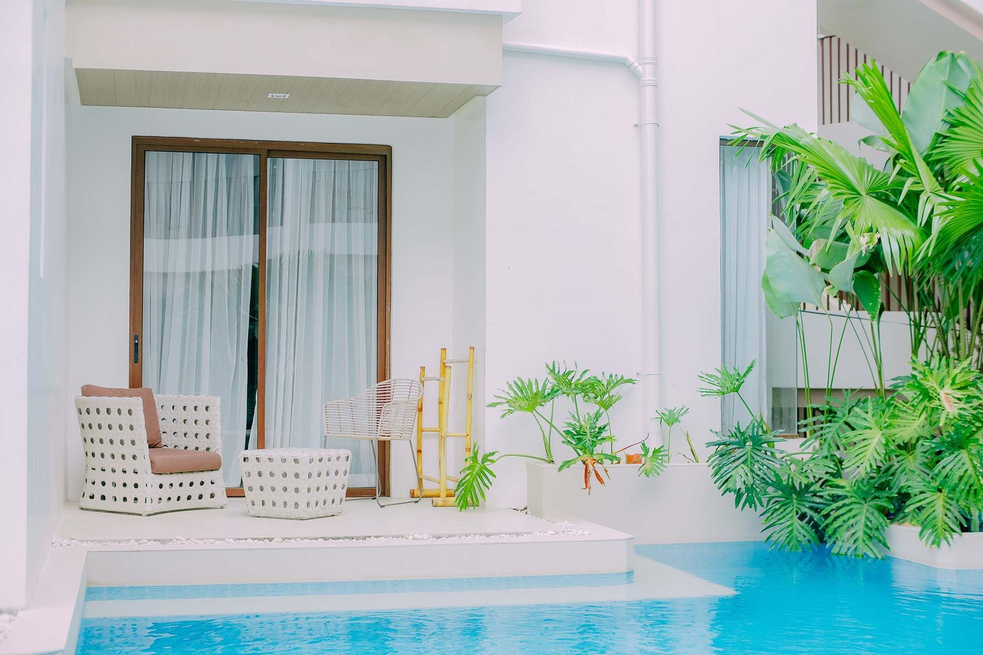 Poolside patio with white seating, sliding doors, and lush green plants beside turquoise water
