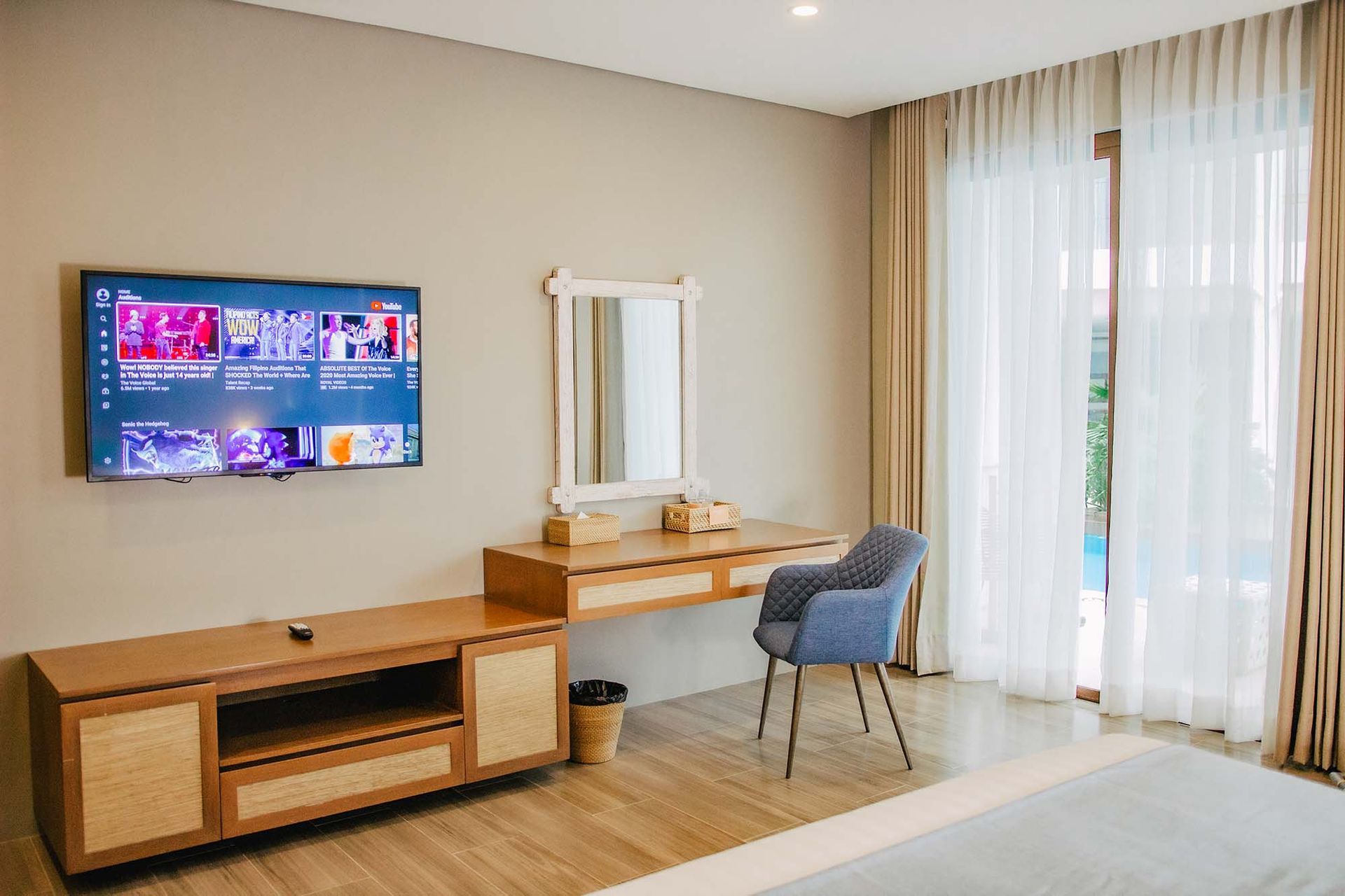 Deluxe room with pool access featuring flat-screen TV, wooden furniture, and blue accent chair by sheer curtains