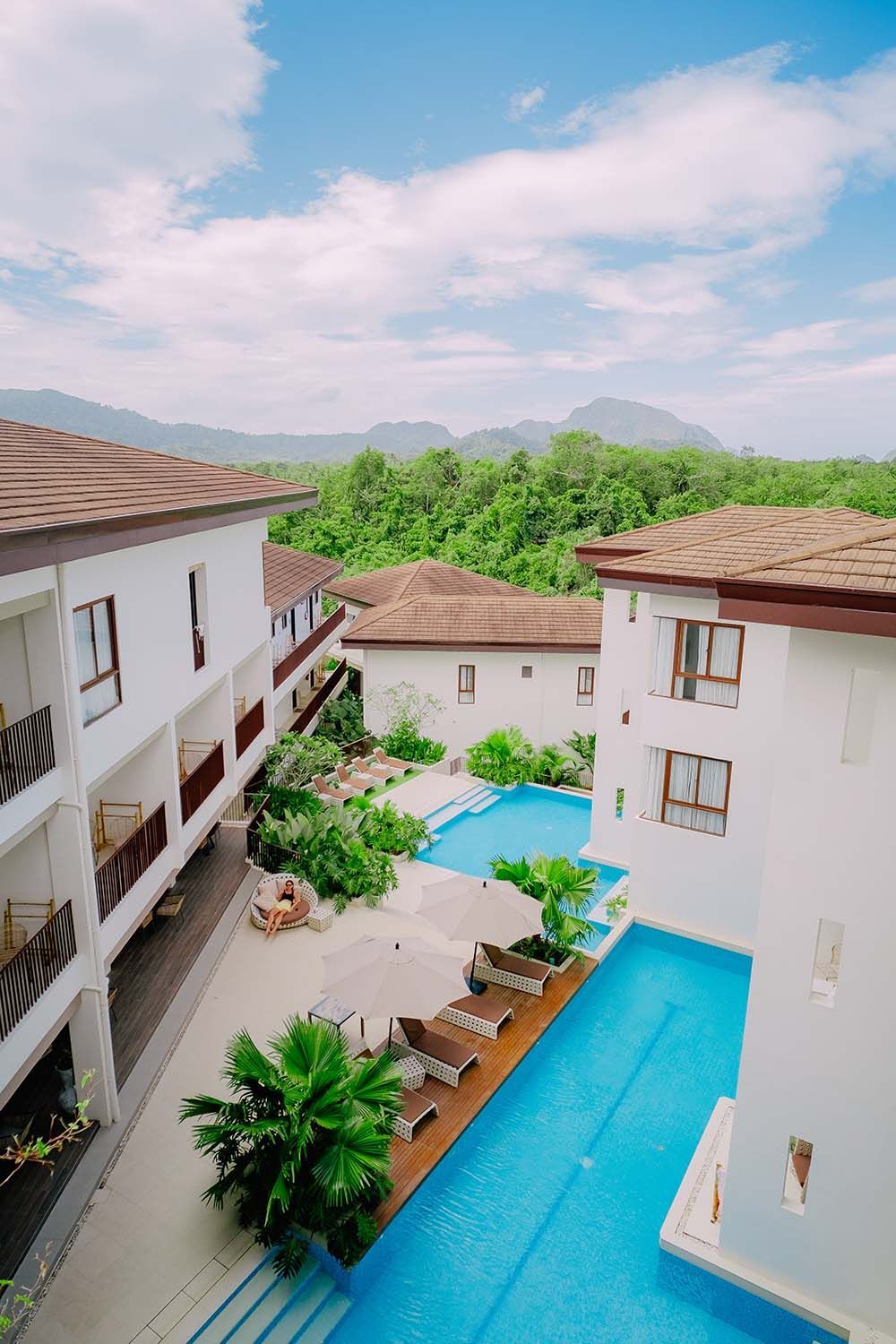 White buildings with brown roofs surround turquoise pools, lush green trees, and mountain backdrop.