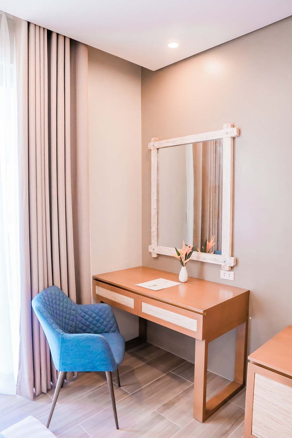 Vanity corner with wooden desk, framed mirror, blue upholstered chair and decorative flower vase