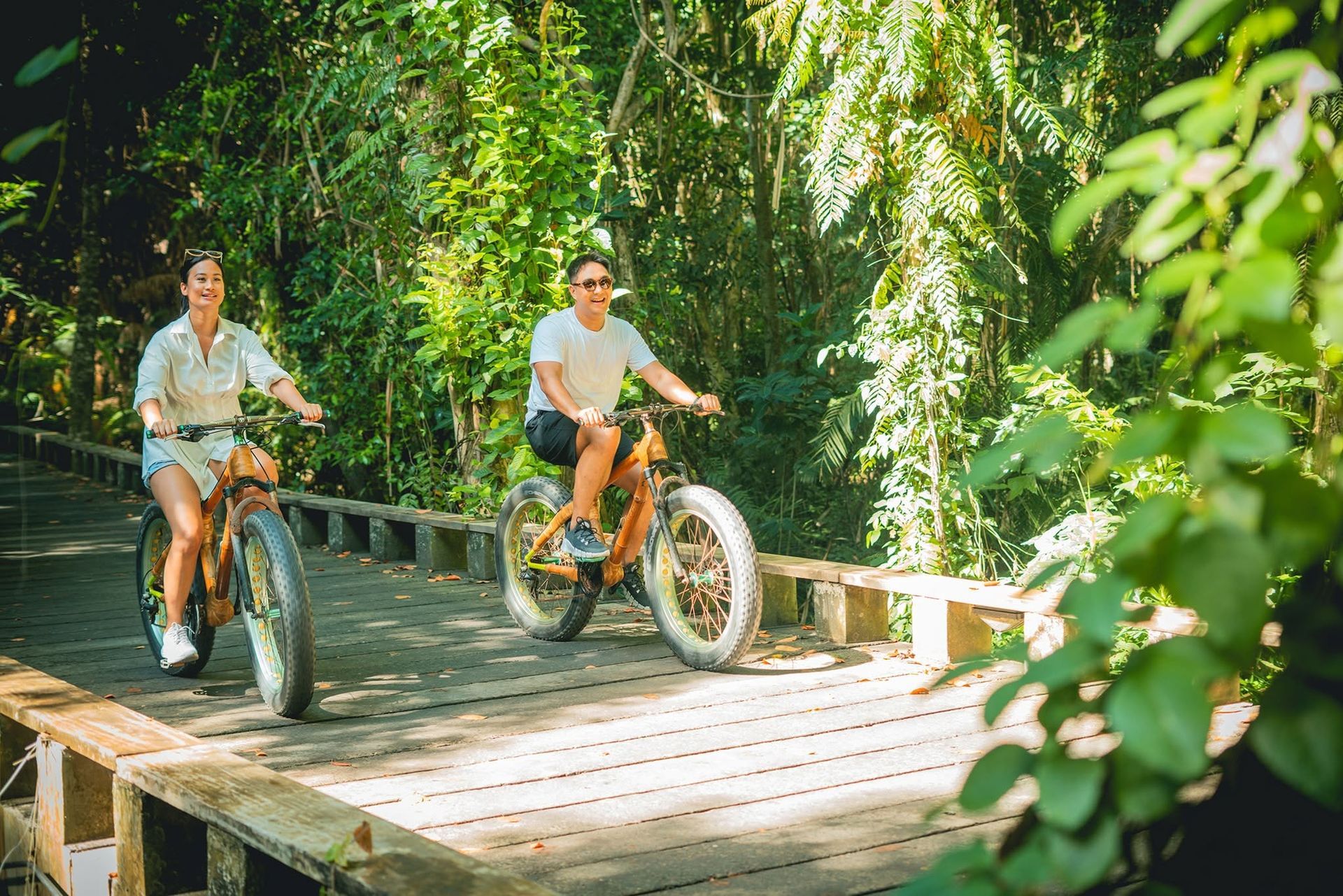 Two people riding bicycles across a wooden bridge surrounded by lush green foliage.