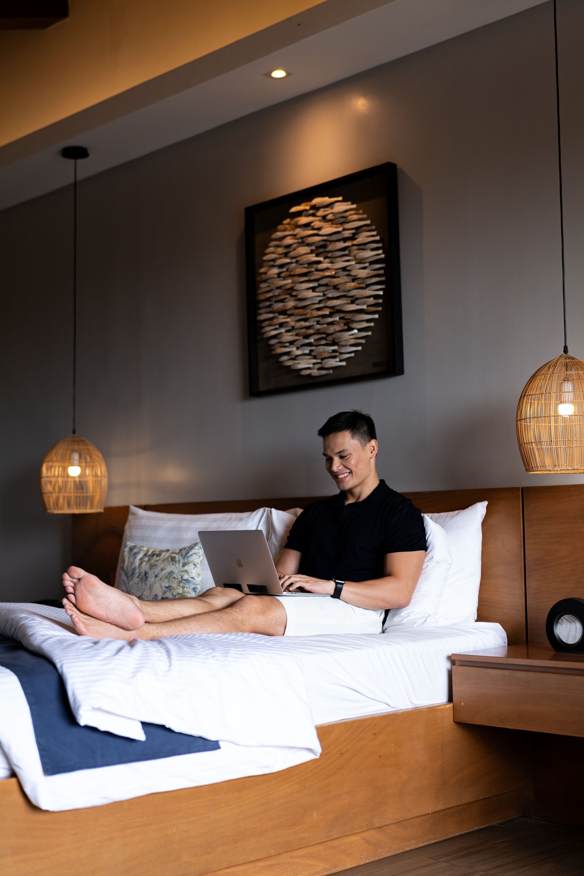 A person sits on a bed in a modern room, smiling while using a laptop with pillows behind them and pendant lights above.