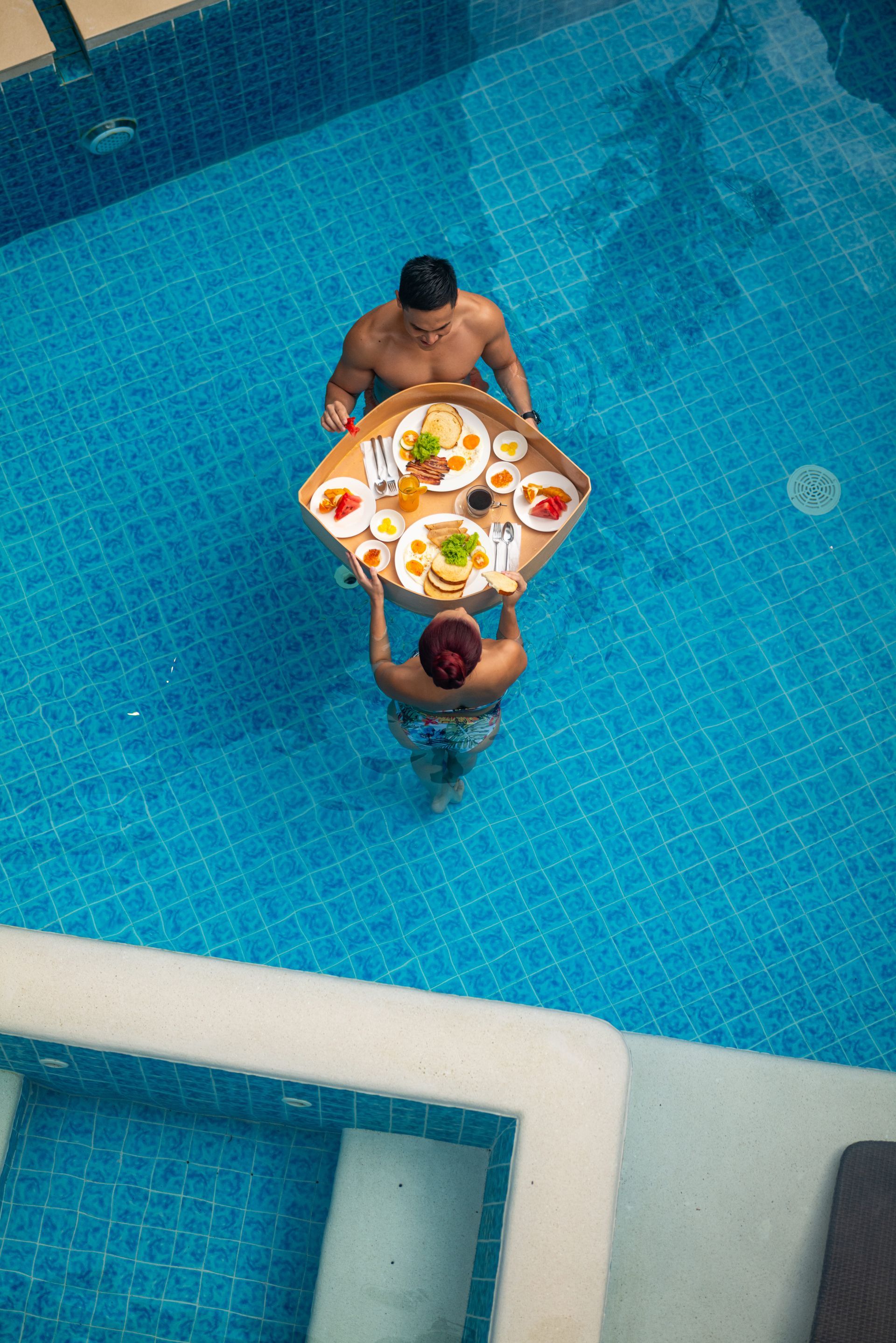 Two people in a blue swimming pool carrying a tray of food between them