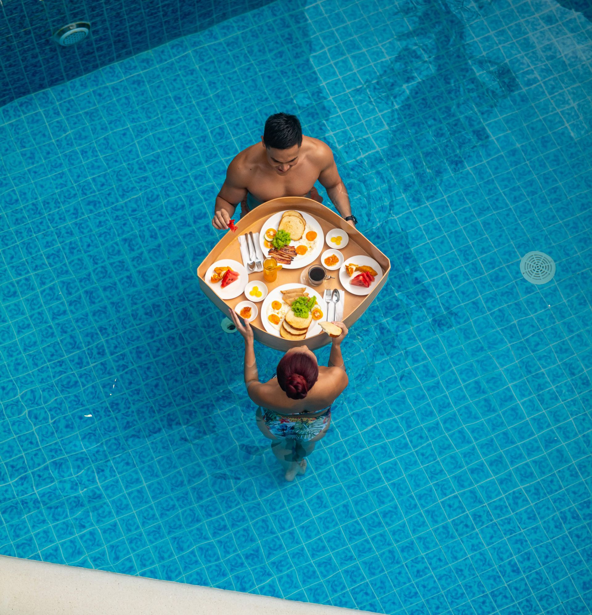 Two people in a blue swimming pool passing a breakfast tray with fruit and drinks
