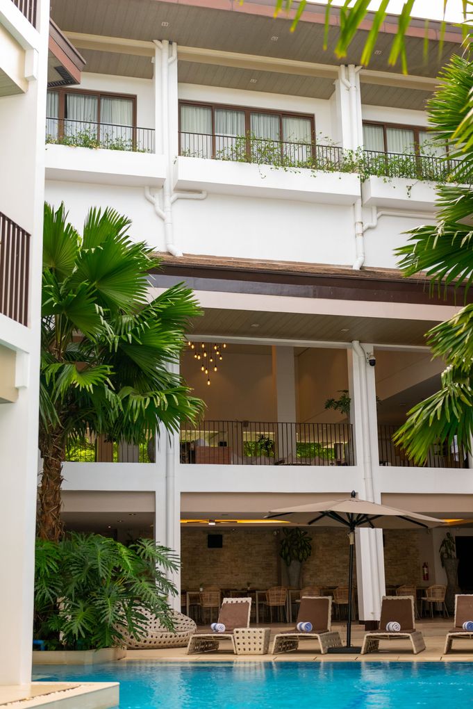 Swimming pool in courtyard next to white building with sliding glass door; lush greenery and blue sky.