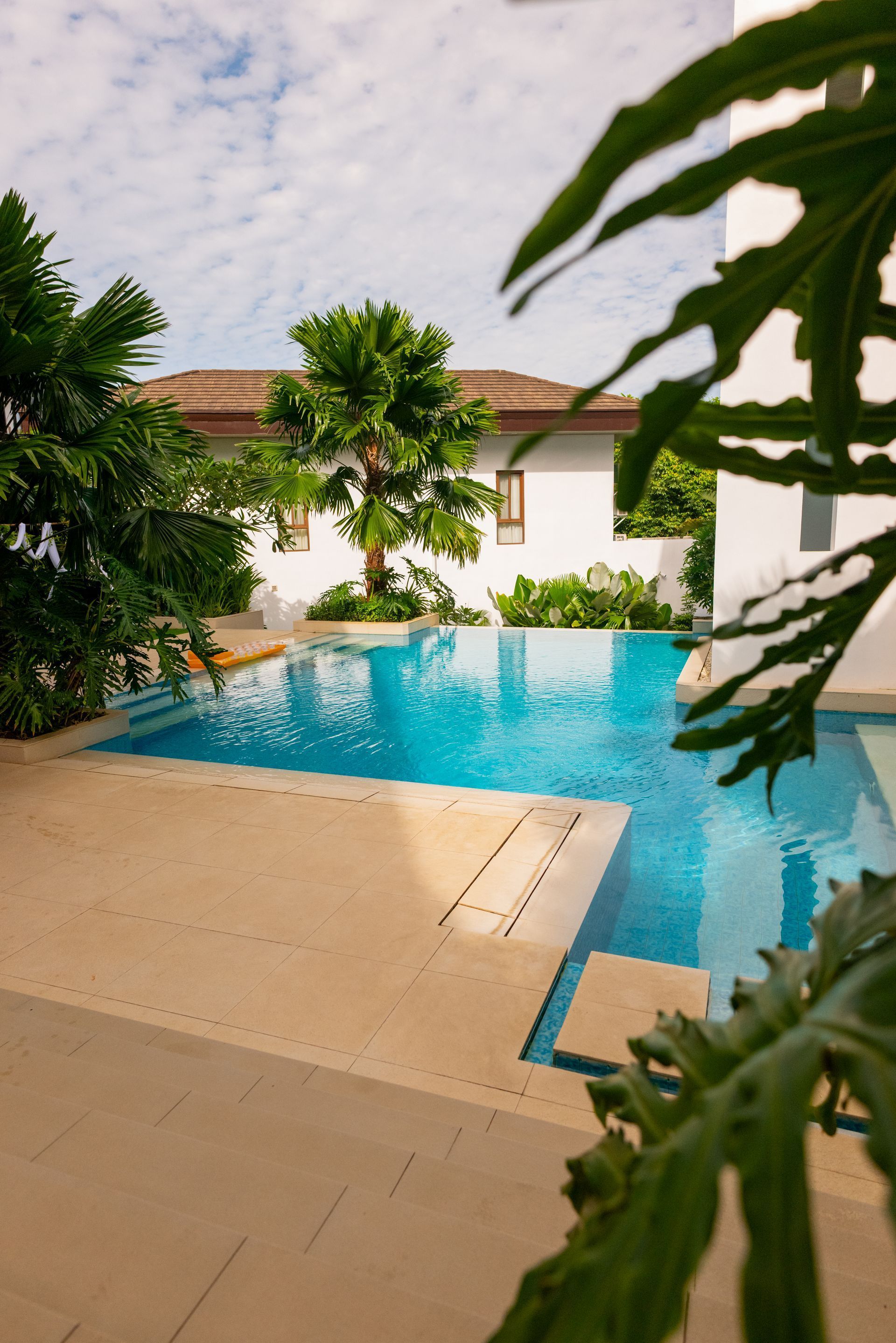Tropical resort pool with turquoise water, tan deck, and white villa under a partly cloudy sky