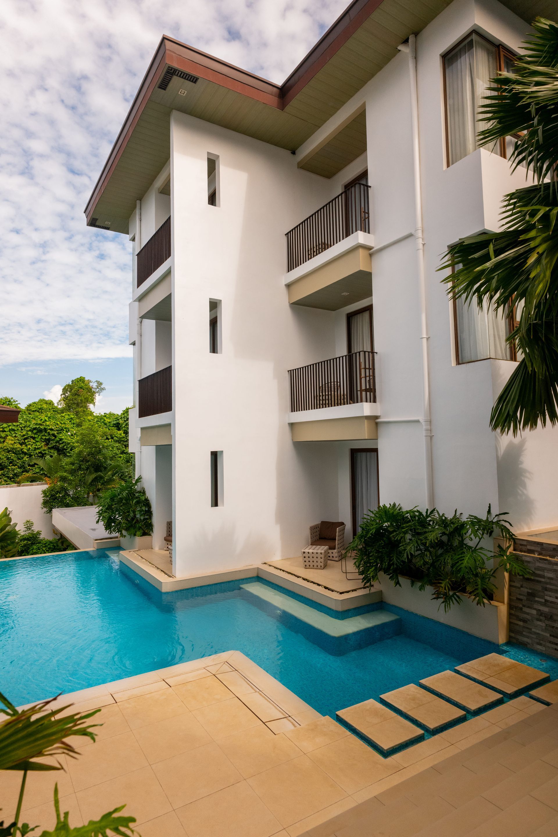 Modern white apartment building beside a turquoise pool with a beige deck and greenery