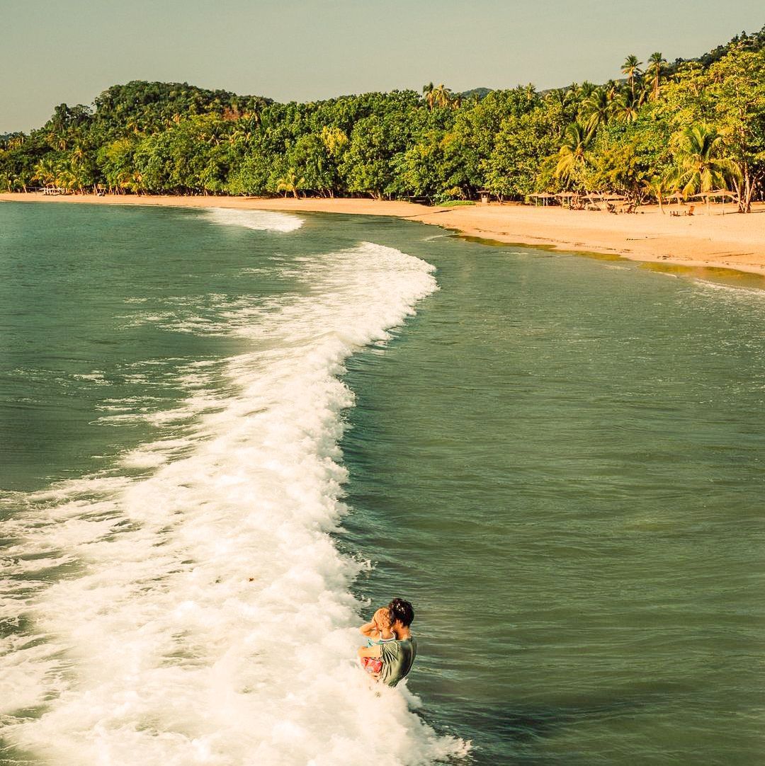 A person stands in the ocean surf near a sandy beach lined with palm trees under a bright sky.