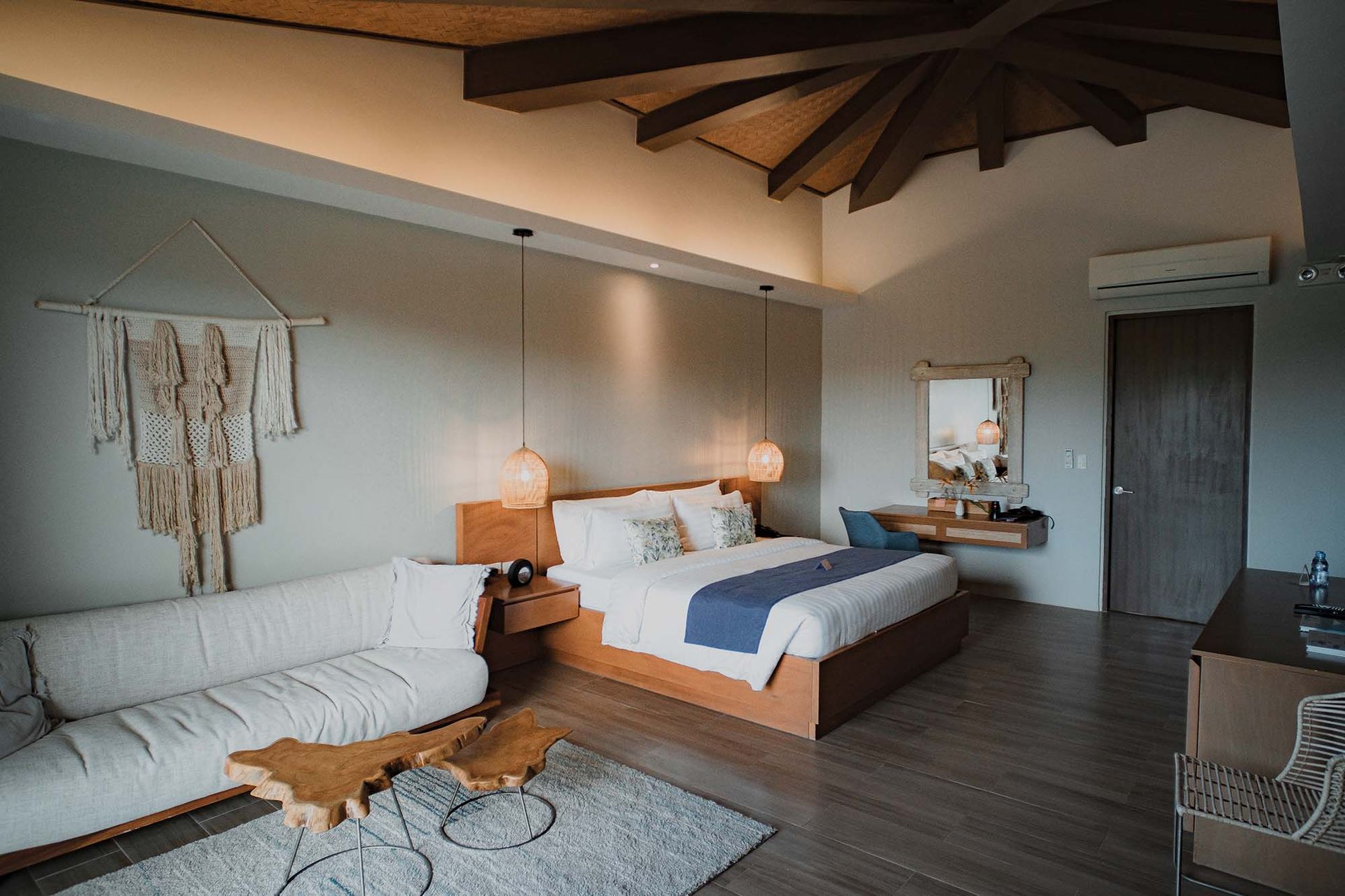 Modern bedroom with wooden beams, white sofa, king bed, and warm hanging lights