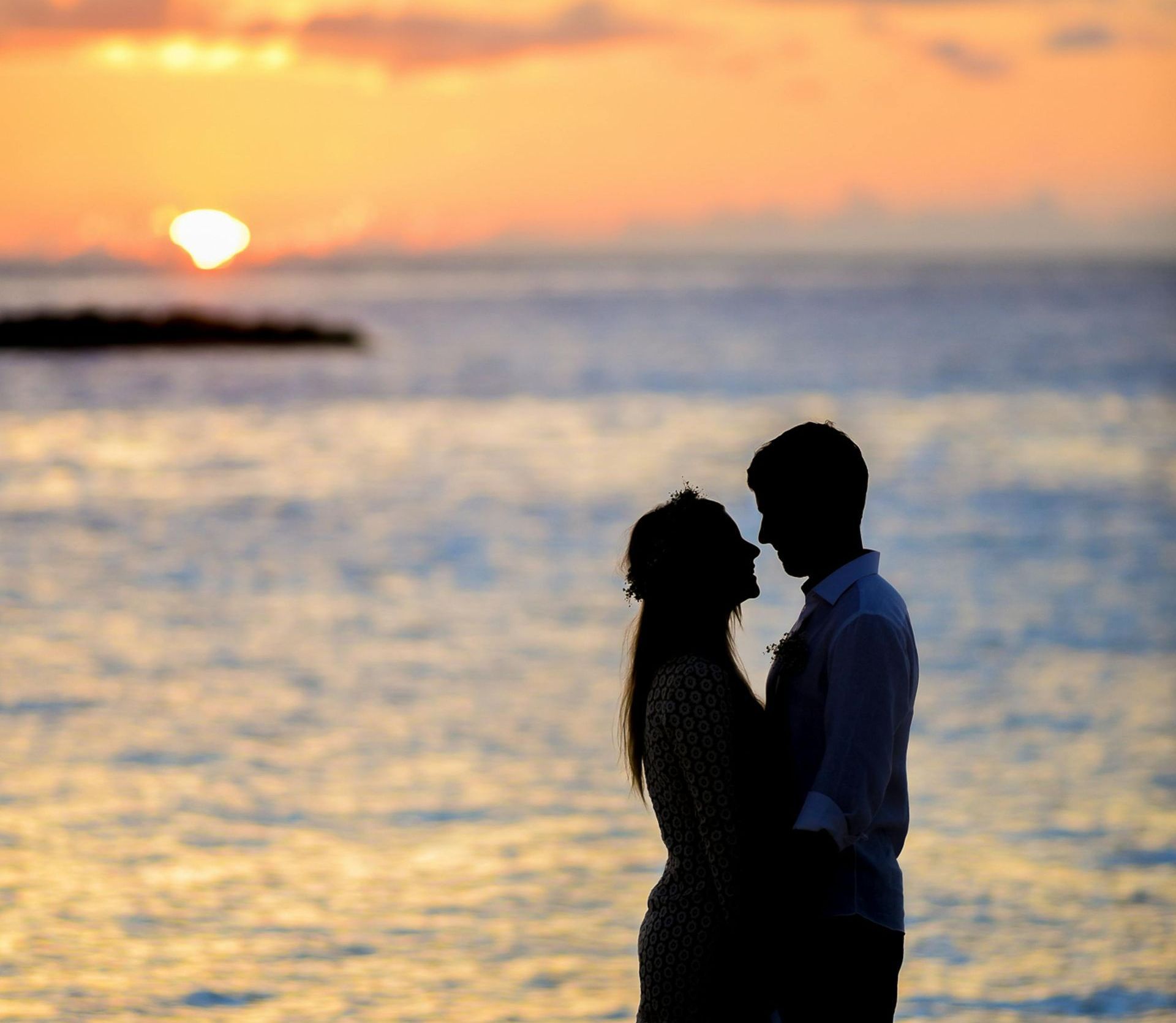 Couple silhouetted embracing on beach at sunset, ocean waves, orange sky.