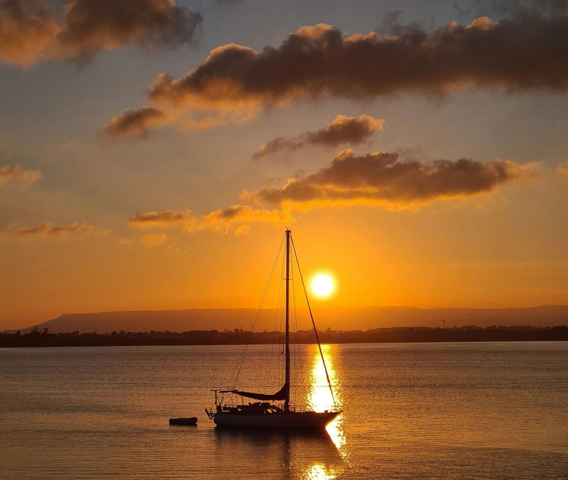 Sailboat silhouetted at sunset on calm water, with orange sky and clouds.