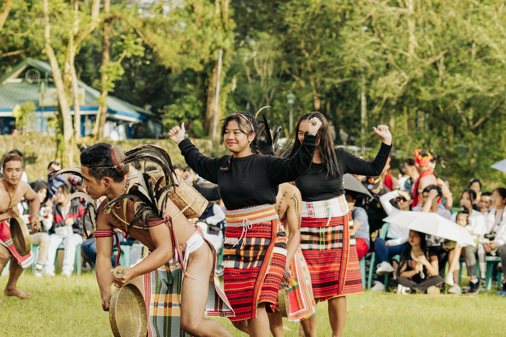 Performers in traditional red skirts dance outdoors before a seated crowd in a park.