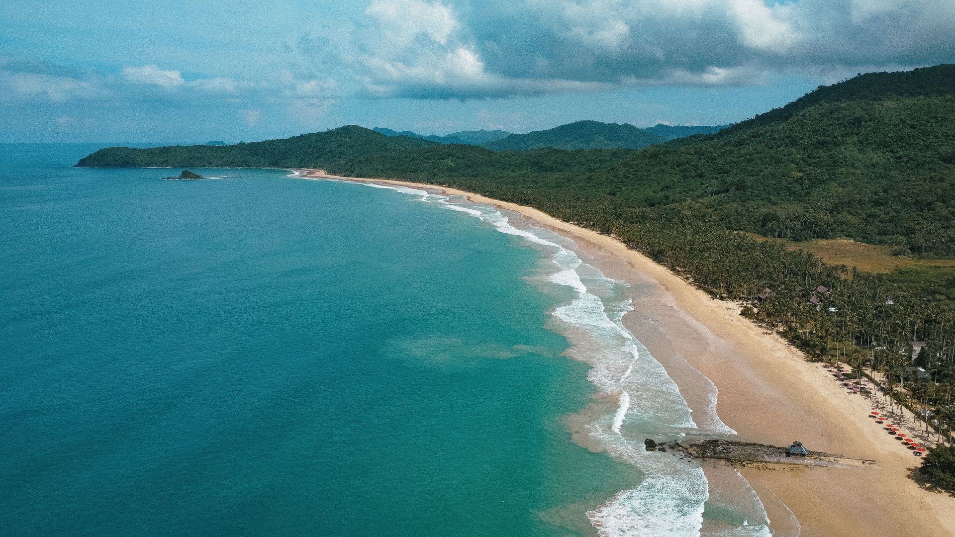 Aerial view of a turquoise beach curving along a green coastal landscape under cloudy skies