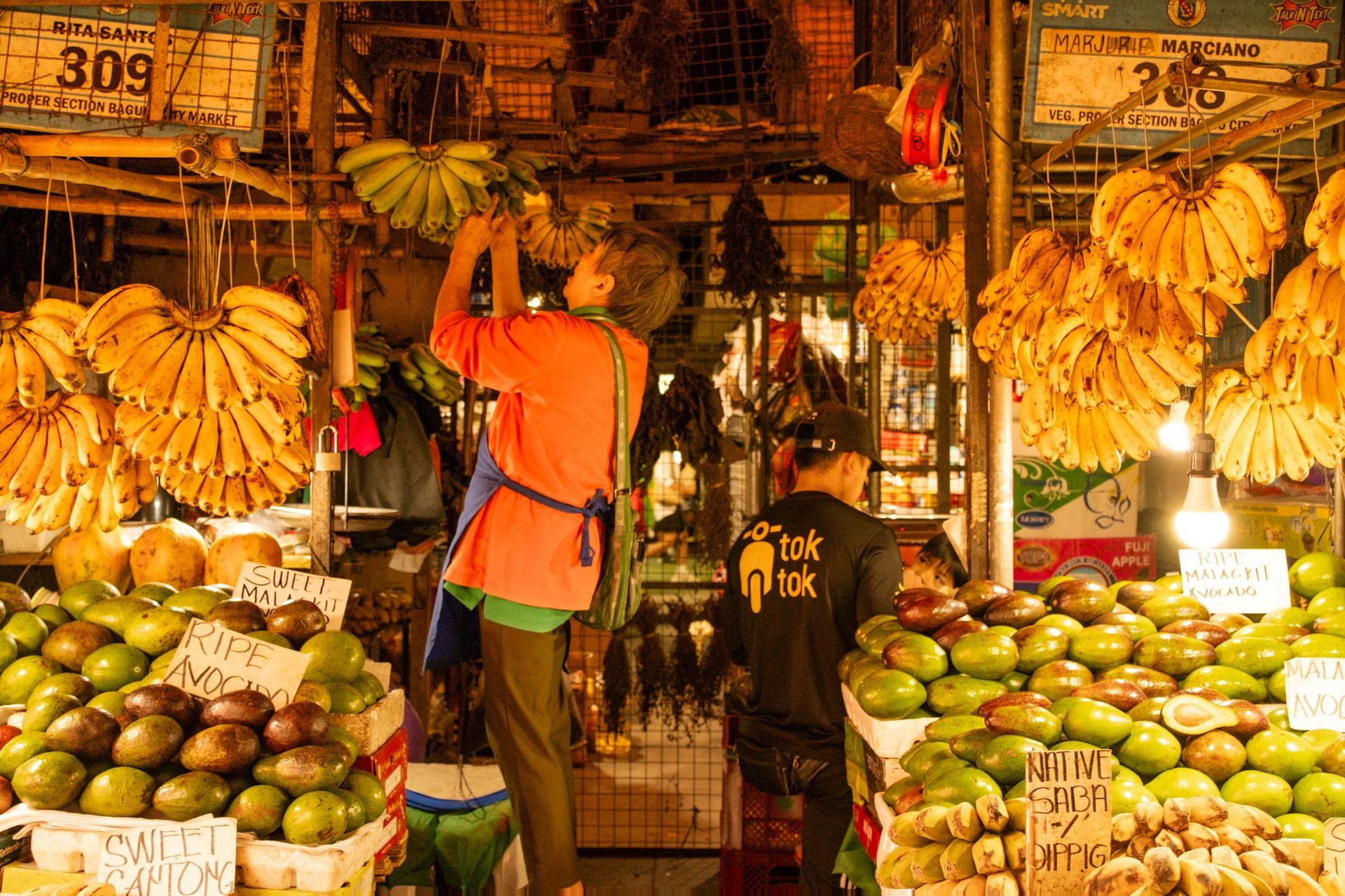Two people in a fruit market browsing bananas, mangoes, and limes under warm lights