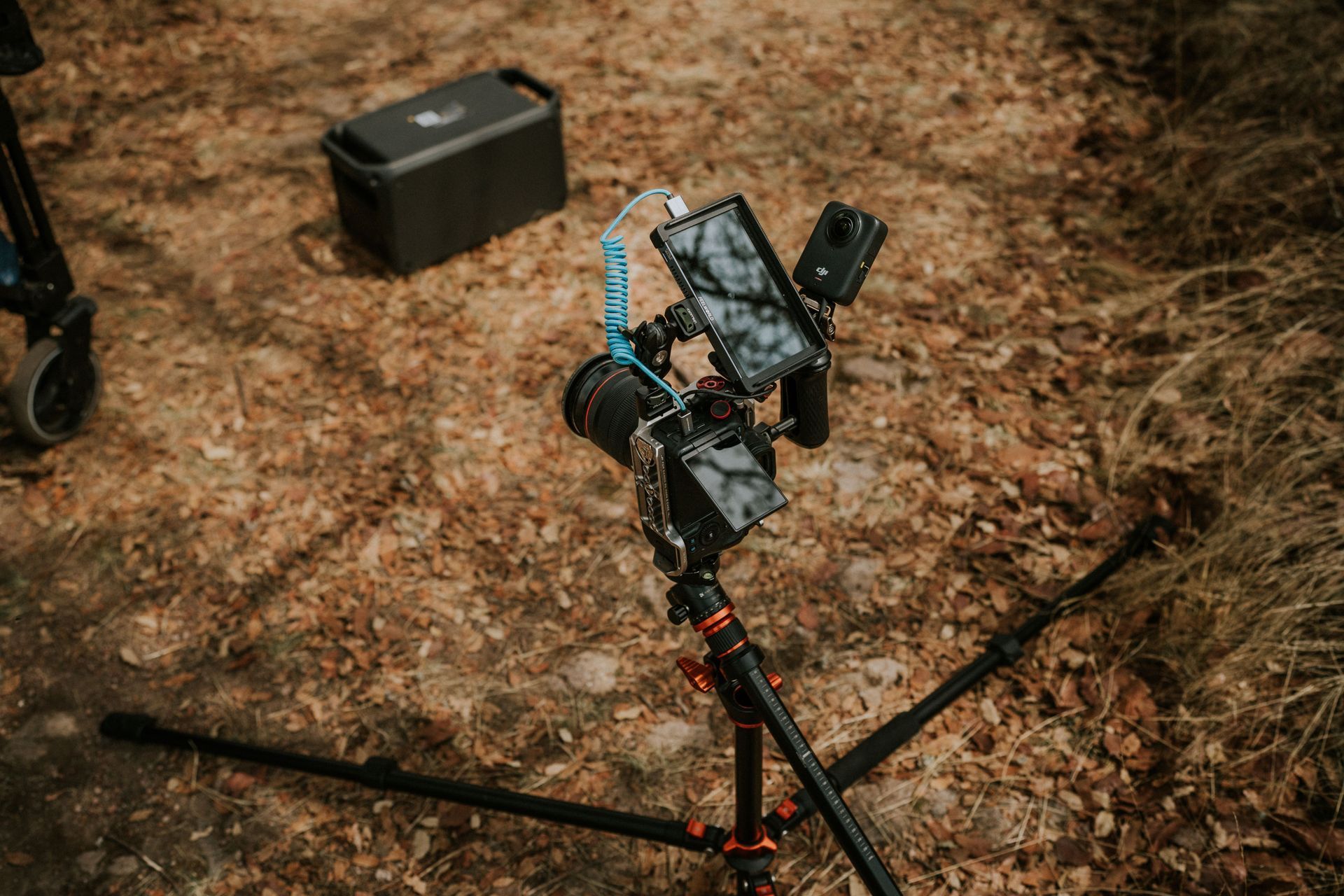 Camera rig on a tripod outdoors in dry grass, with a small equipment case nearby.