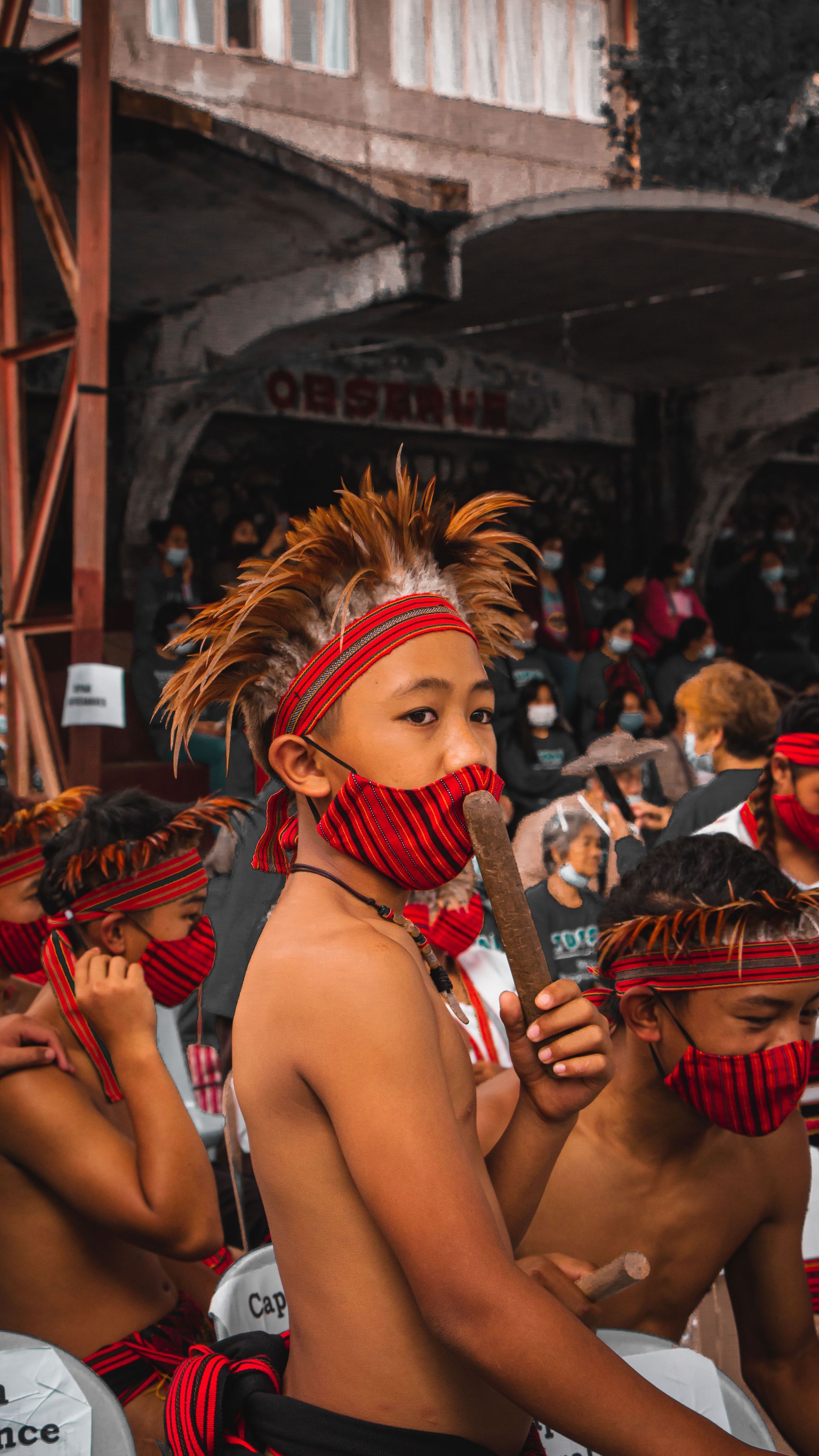 Masked dancers in red feathered headdresses and face coverings during a crowded street festival