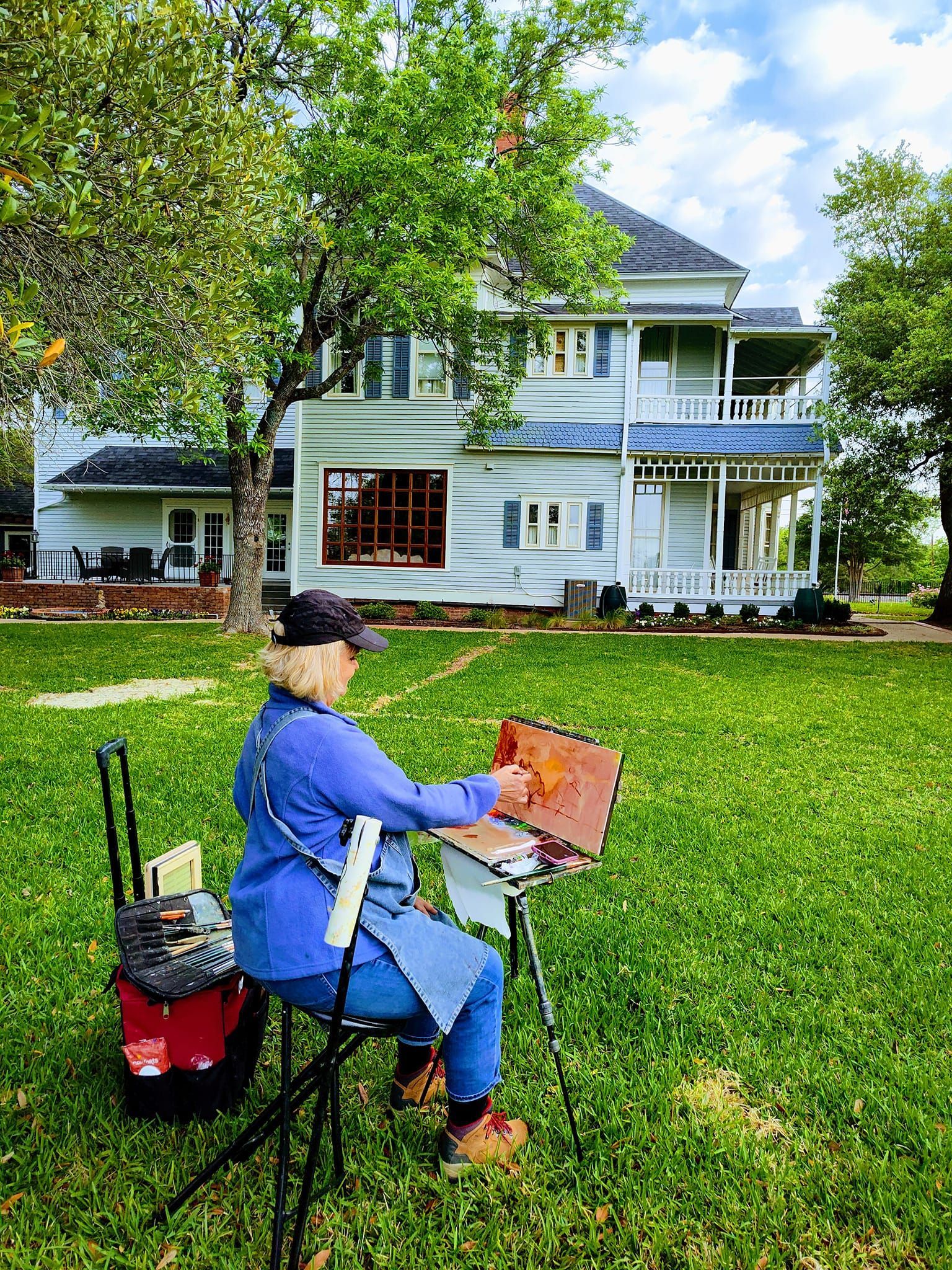A woman is sitting in the grass painting a picture of a house.