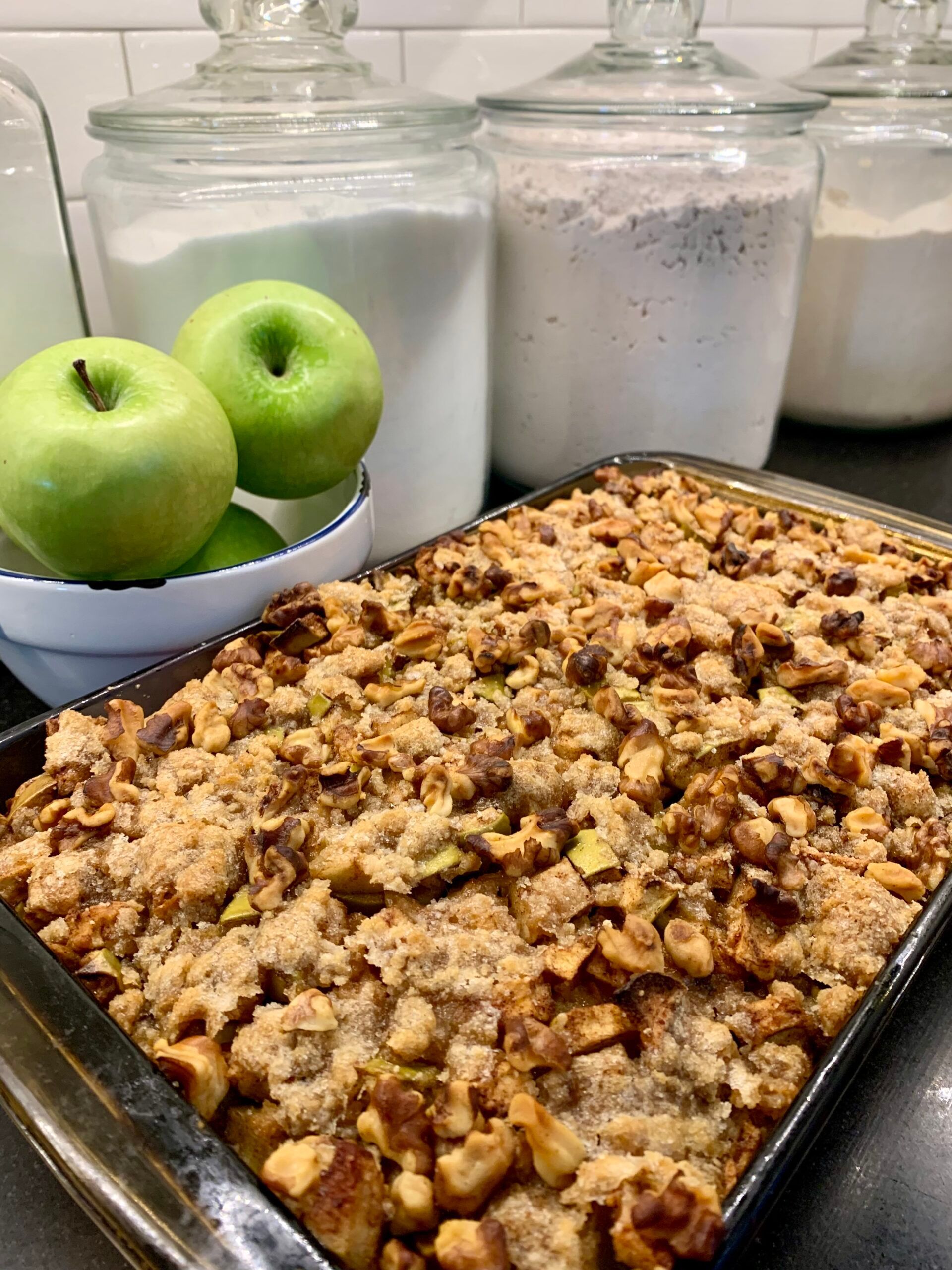 Apple crisp in a baking pan, with apples in a bowl and canisters of ingredients in background.