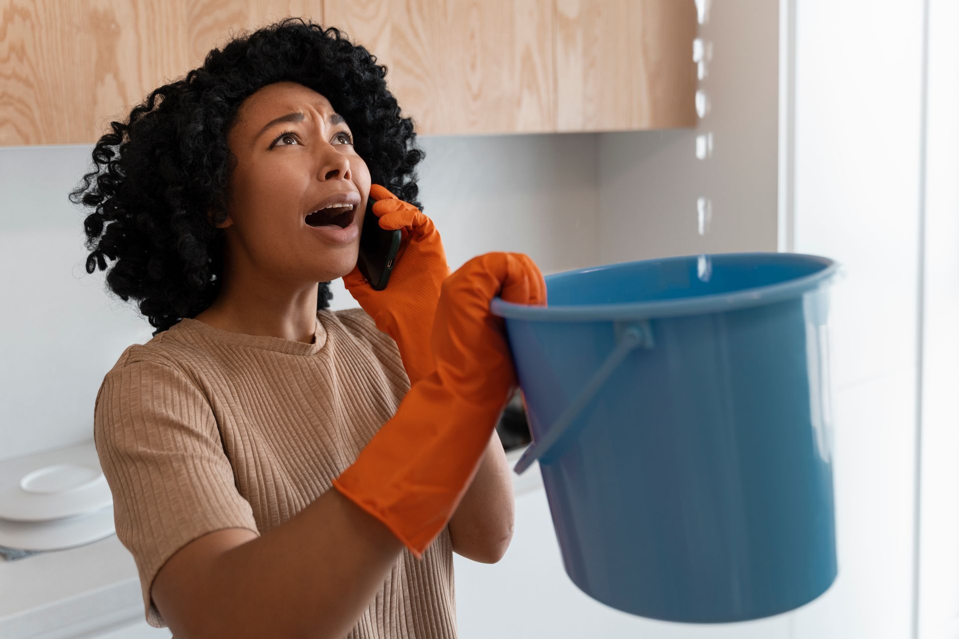 Person in orange gloves holding a blue bucket and talking on a phone in a kitchen