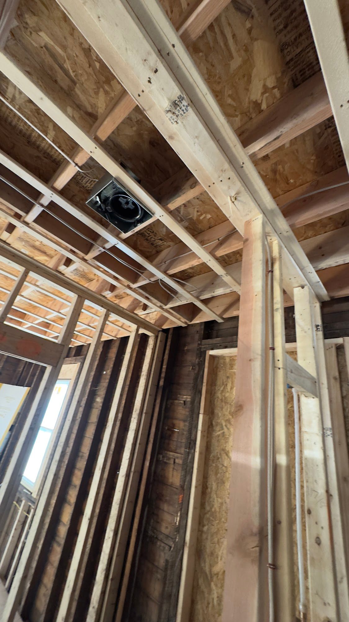 Interior view of a building under construction, showing framing, ceiling beams, and a vent. Wooden structure.