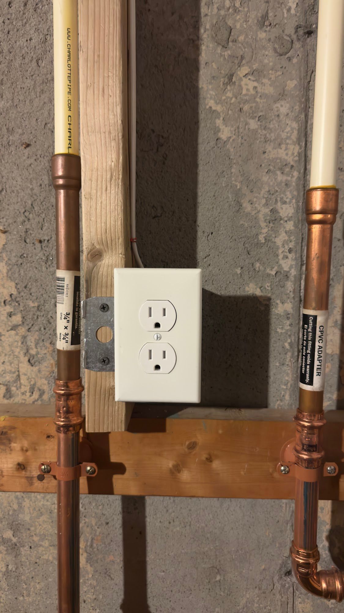 Electrical outlet mounted on wooden studs between copper pipes and PVC pipes against a concrete wall.