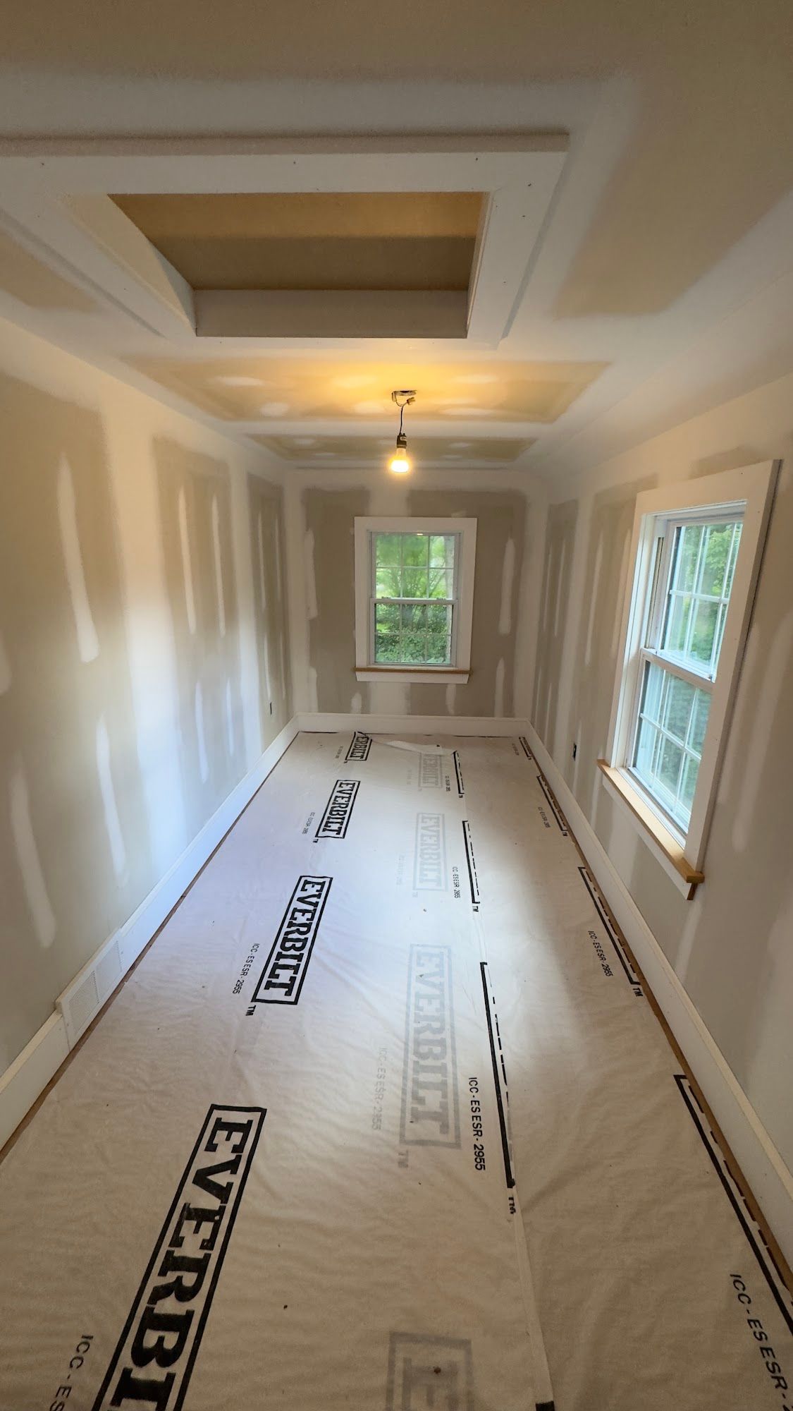 A room under construction with drywall and protective flooring. Windows and a ceiling opening are visible.